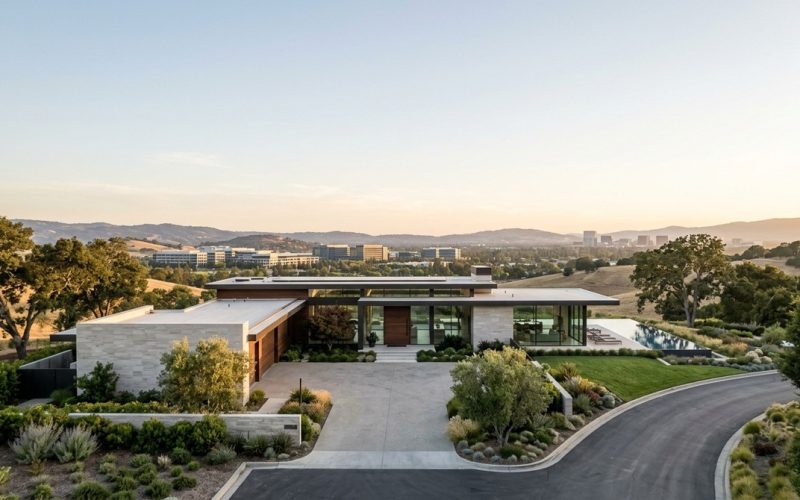 Modern luxury home with an infinity pool overlooking a valley and distant city buildings at sunset.