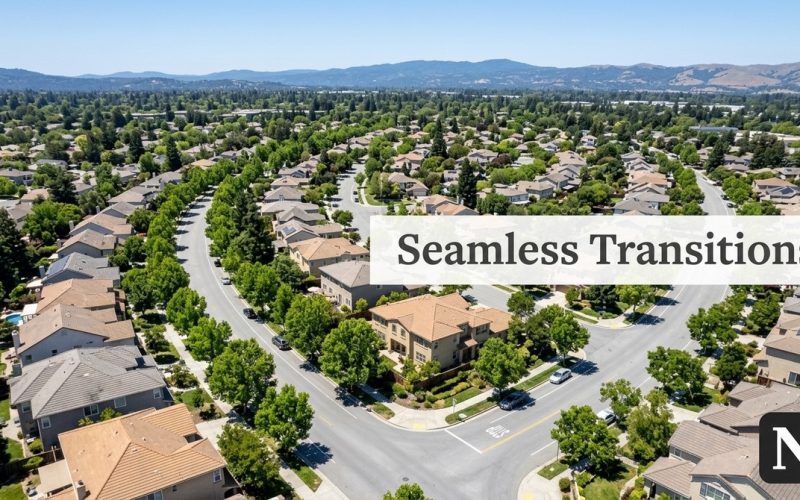 Aerial view of a suburban neighborhood with many houses, green trees, and winding roads. Mountains in distance.